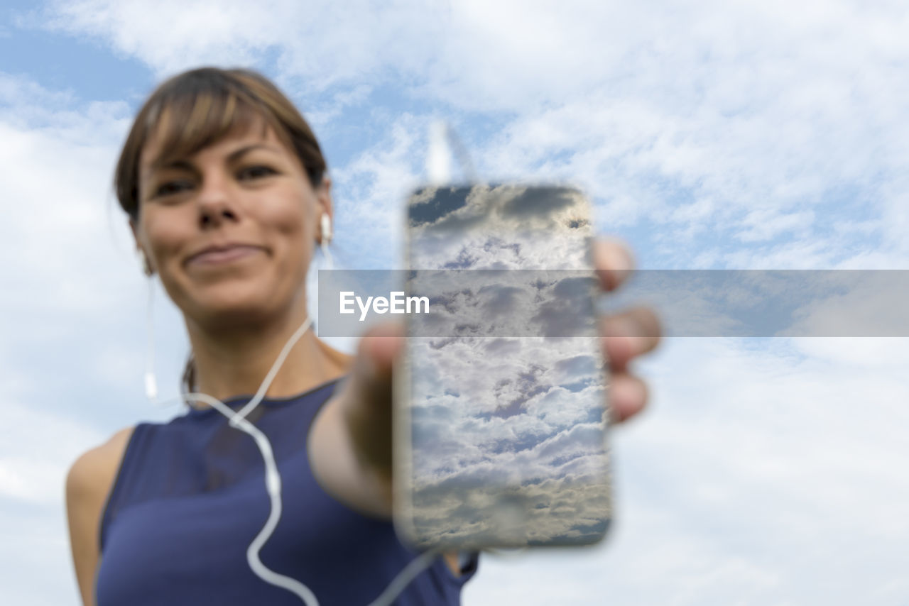 Portrait of smiling young woman showing mobile phone with clouds reflection