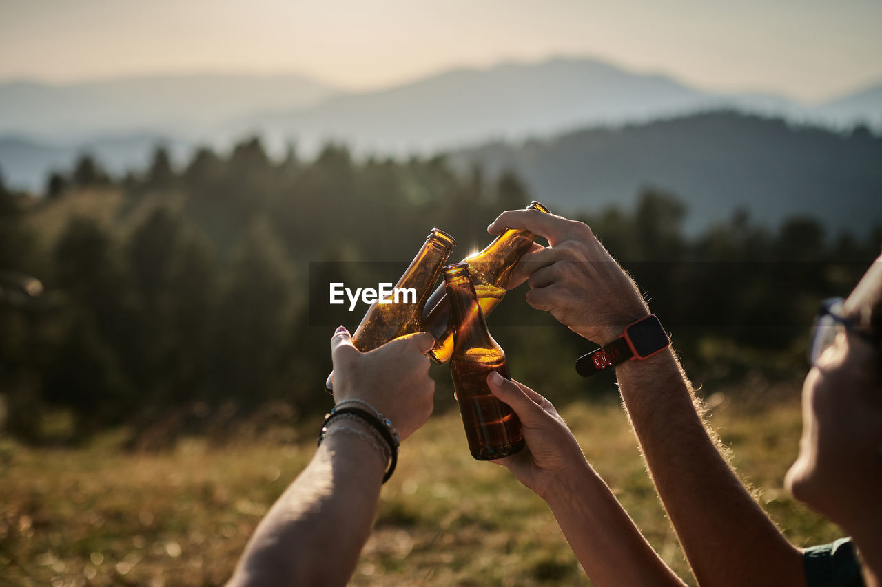 Company of unrecognizable friends relaxing in highland area and clinking bottles of beer while enjoying sunset in summer during weekend