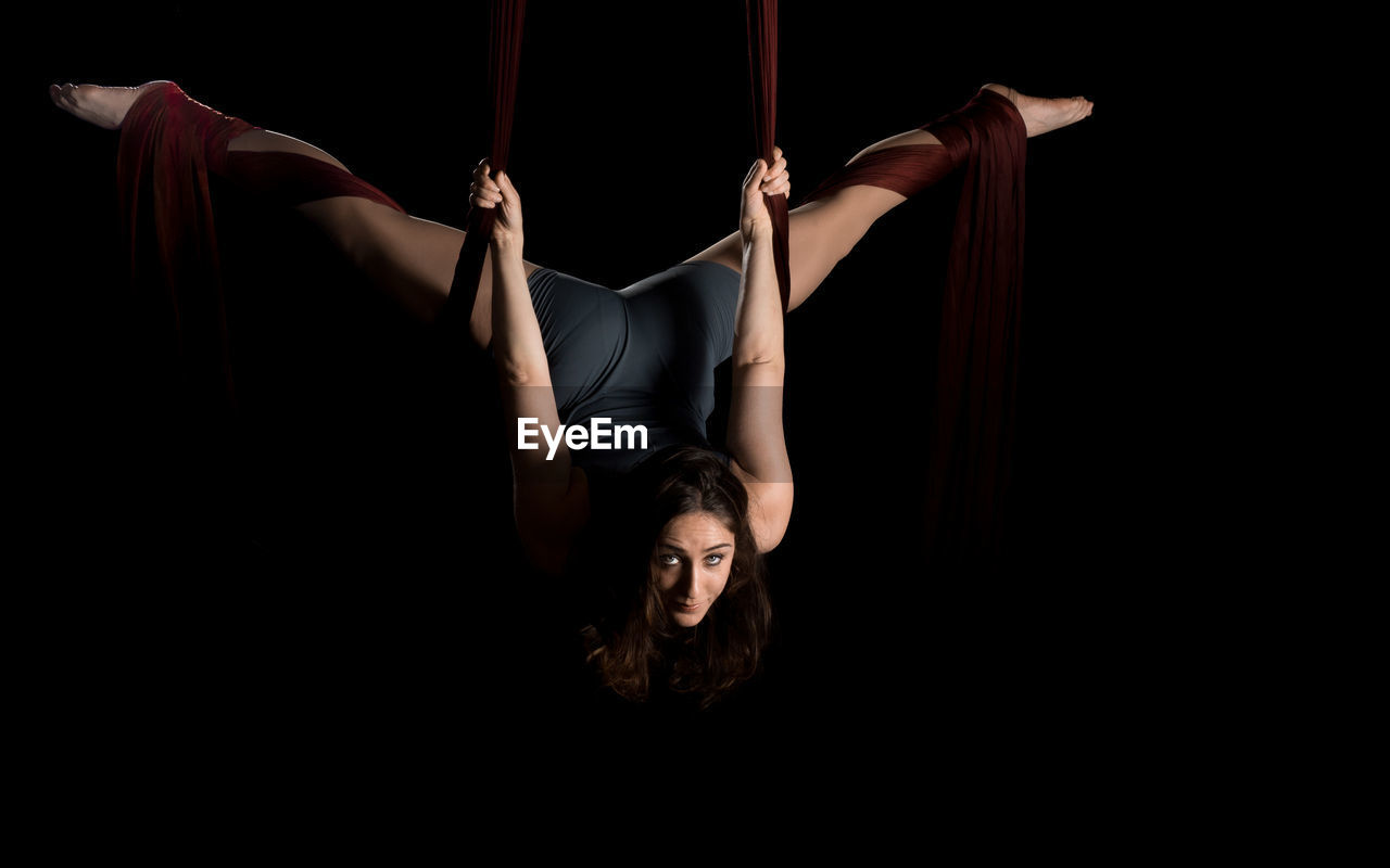 Young woman hanging on fabric while dancing against black background