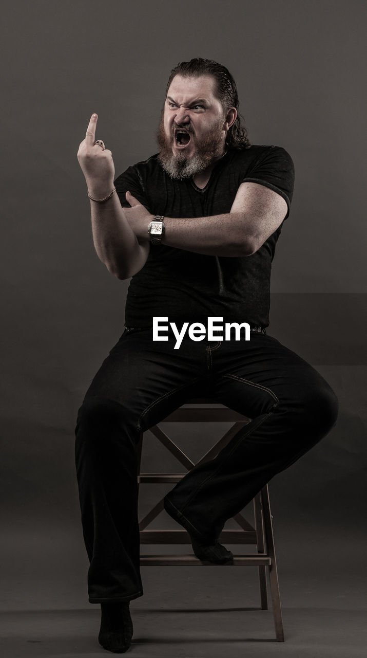 Mid adult man gesturing obscene gesture while sitting against black background