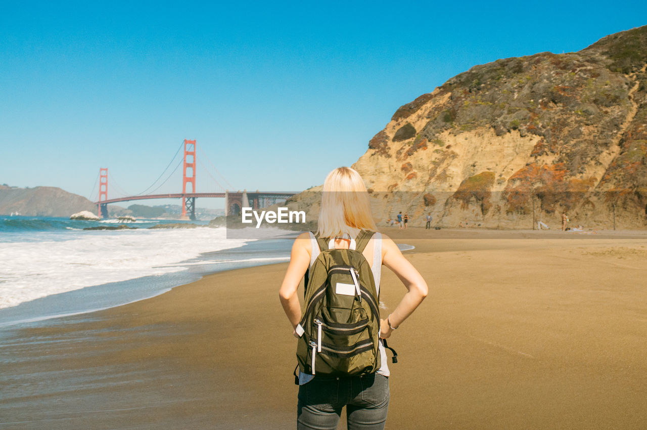 Rear view of woman standing on shore with golden gate bridge against clear sky