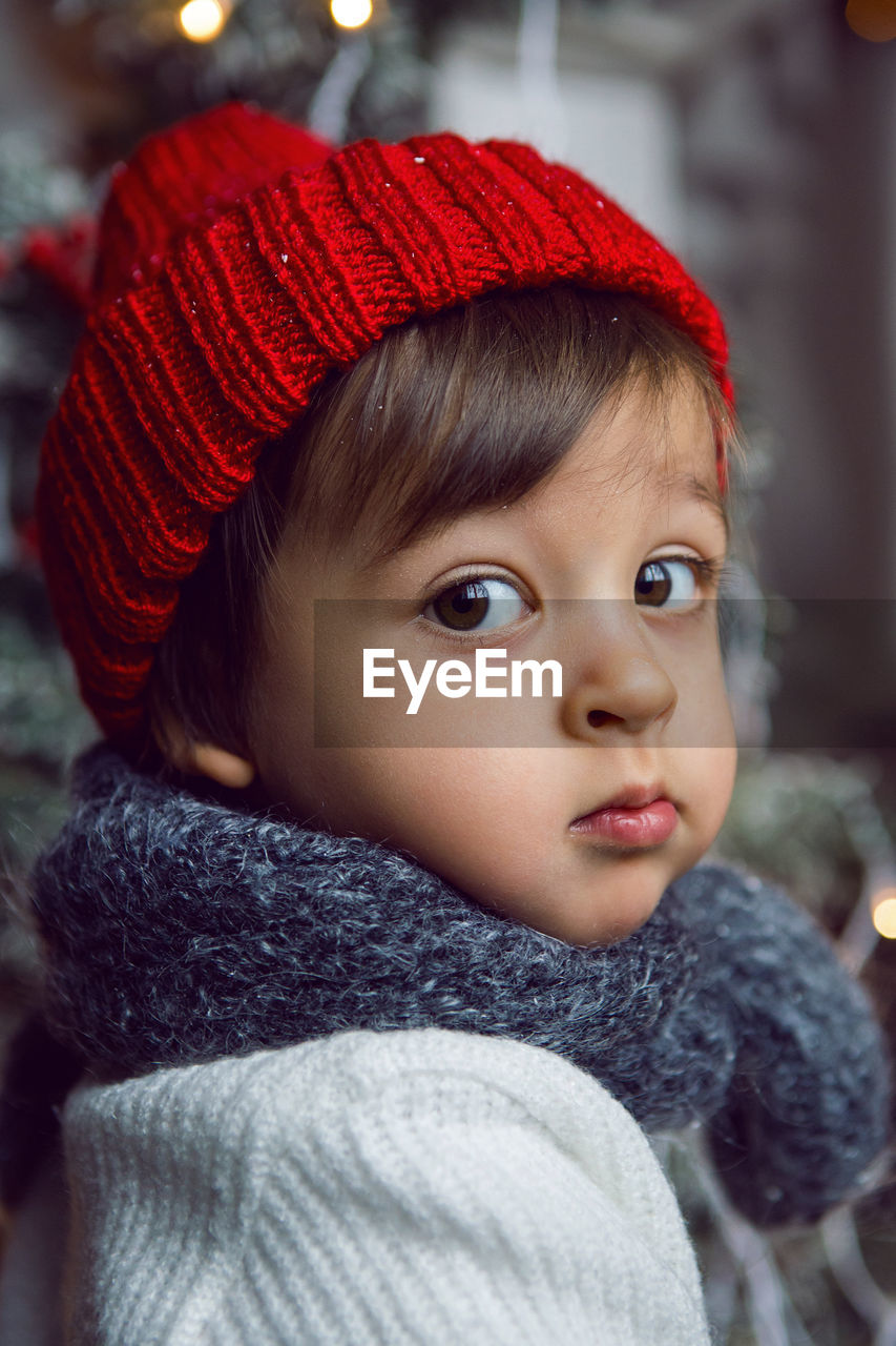 Child in a white sweater and a red knitted hat sits on the steps with snow at home at christmas