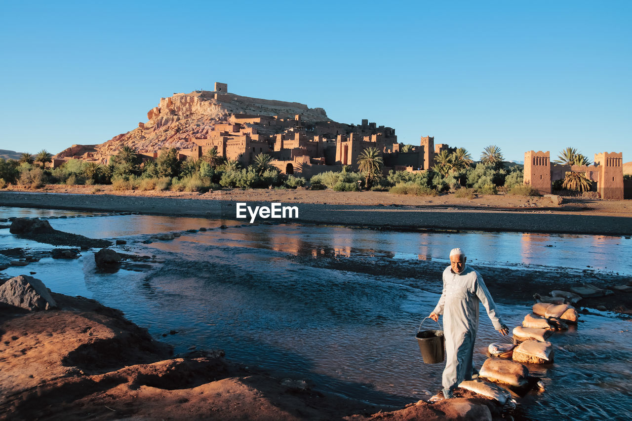 Man carrying bucket while crossing river against old ruins