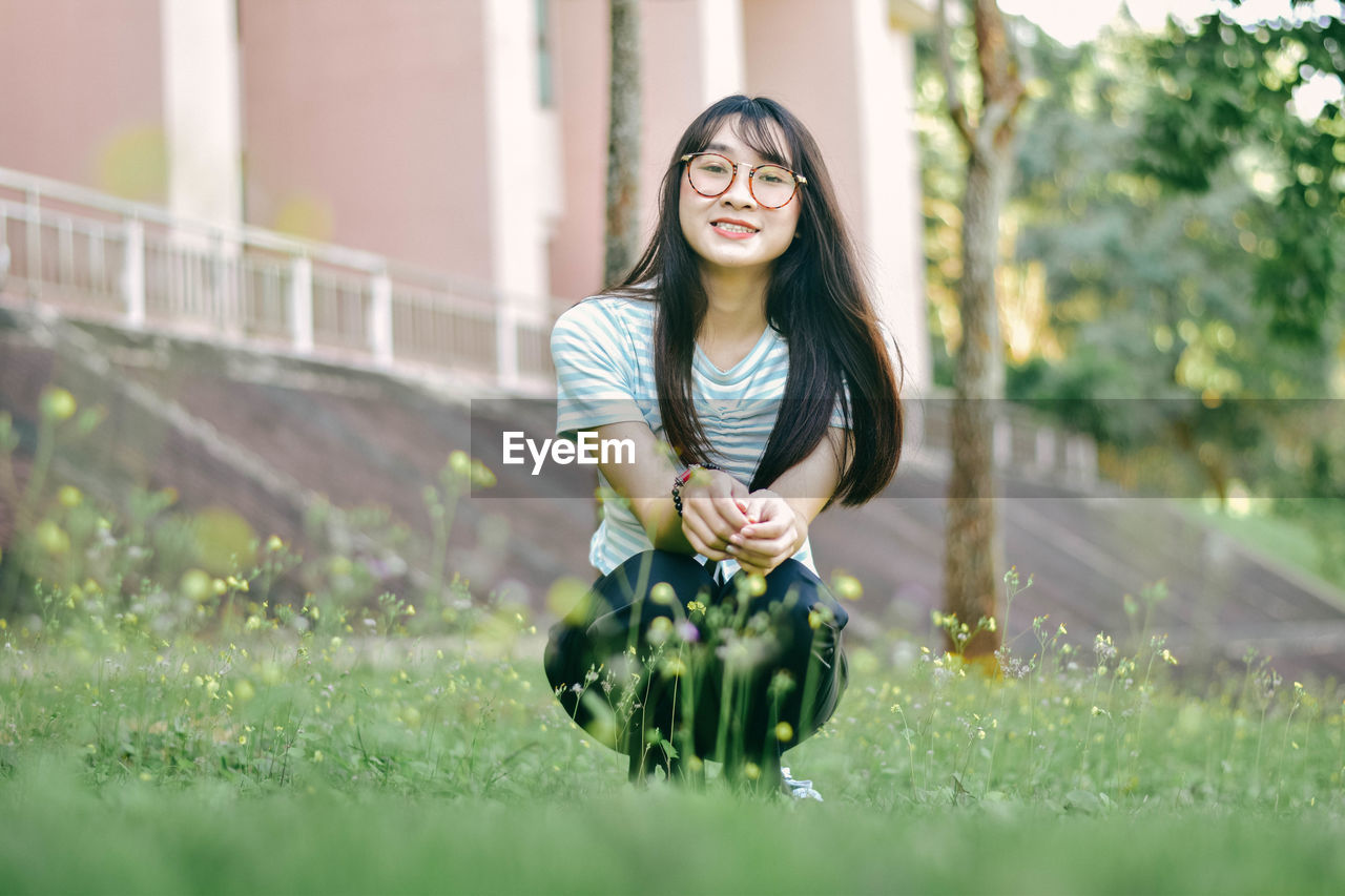 Portrait of cheerful young woman crouching on grass against building at park