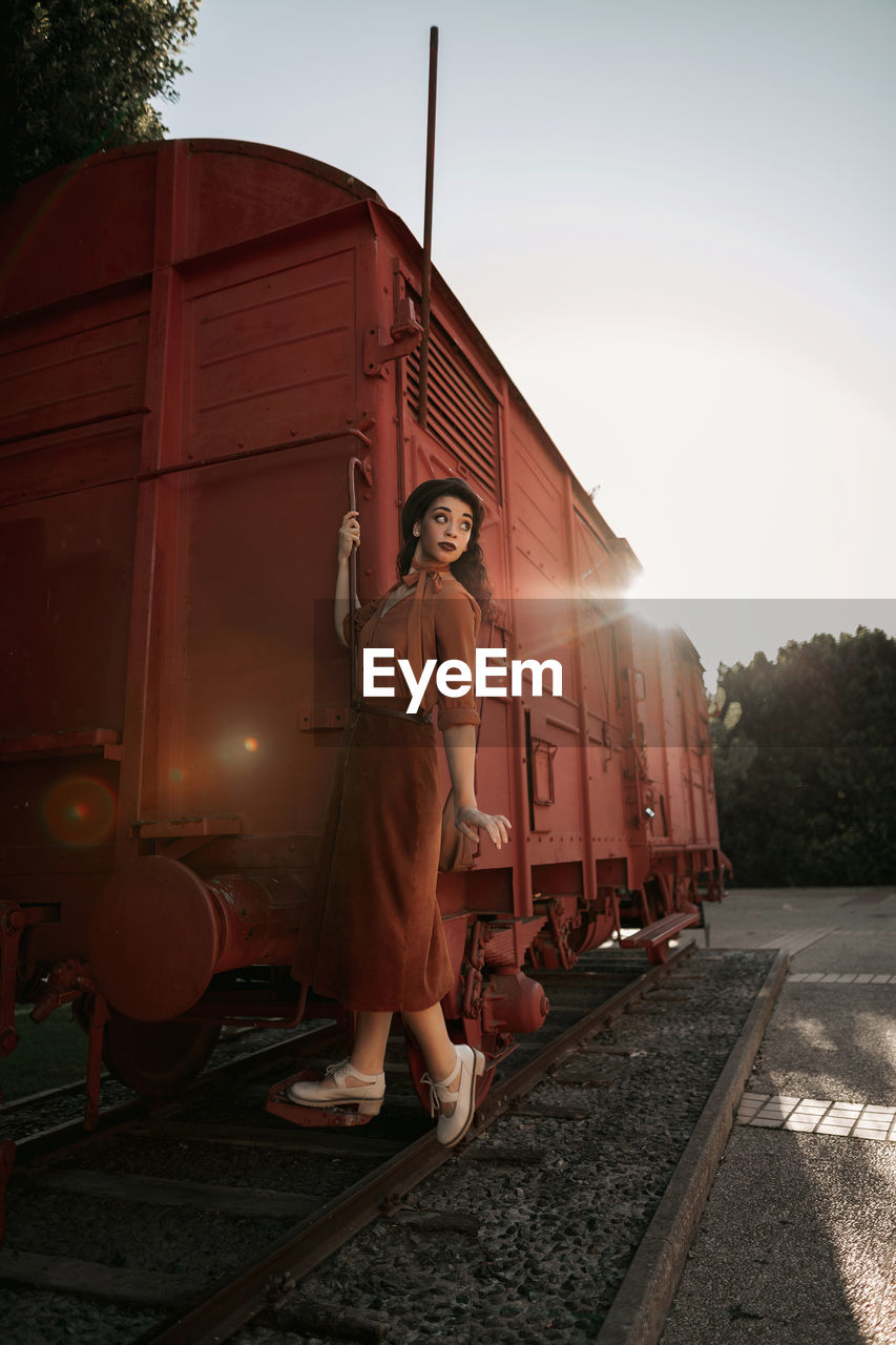 Young female with dark curly hair in beret wearing terracotta clothes in vintage style in back lit standing on step of car train painted in terracotta color and holding handrail while looking away flirty