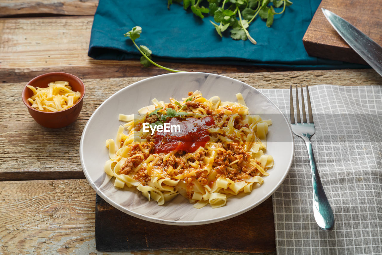 Pasta bolognese garnished with greens and cheese in a plate on a wooden table next to a fork