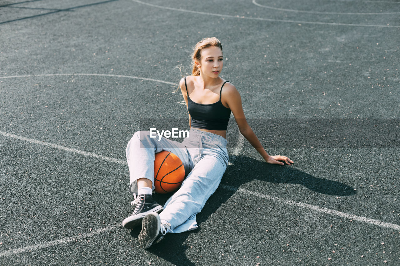 A basketball player is sitting on a sports field with a basketball and looks away. sport