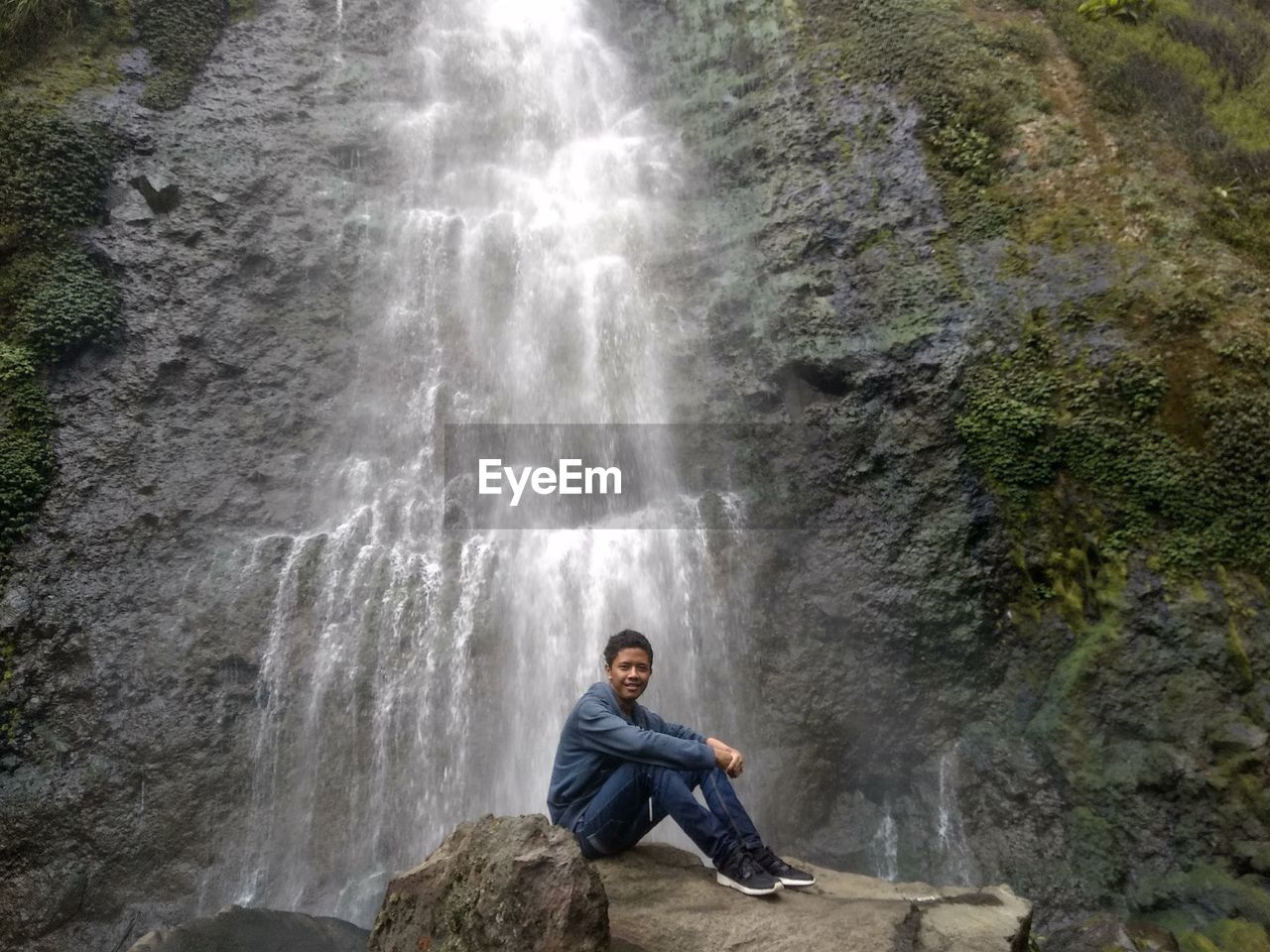 MAN SITTING ON ROCK AT WATERFALL