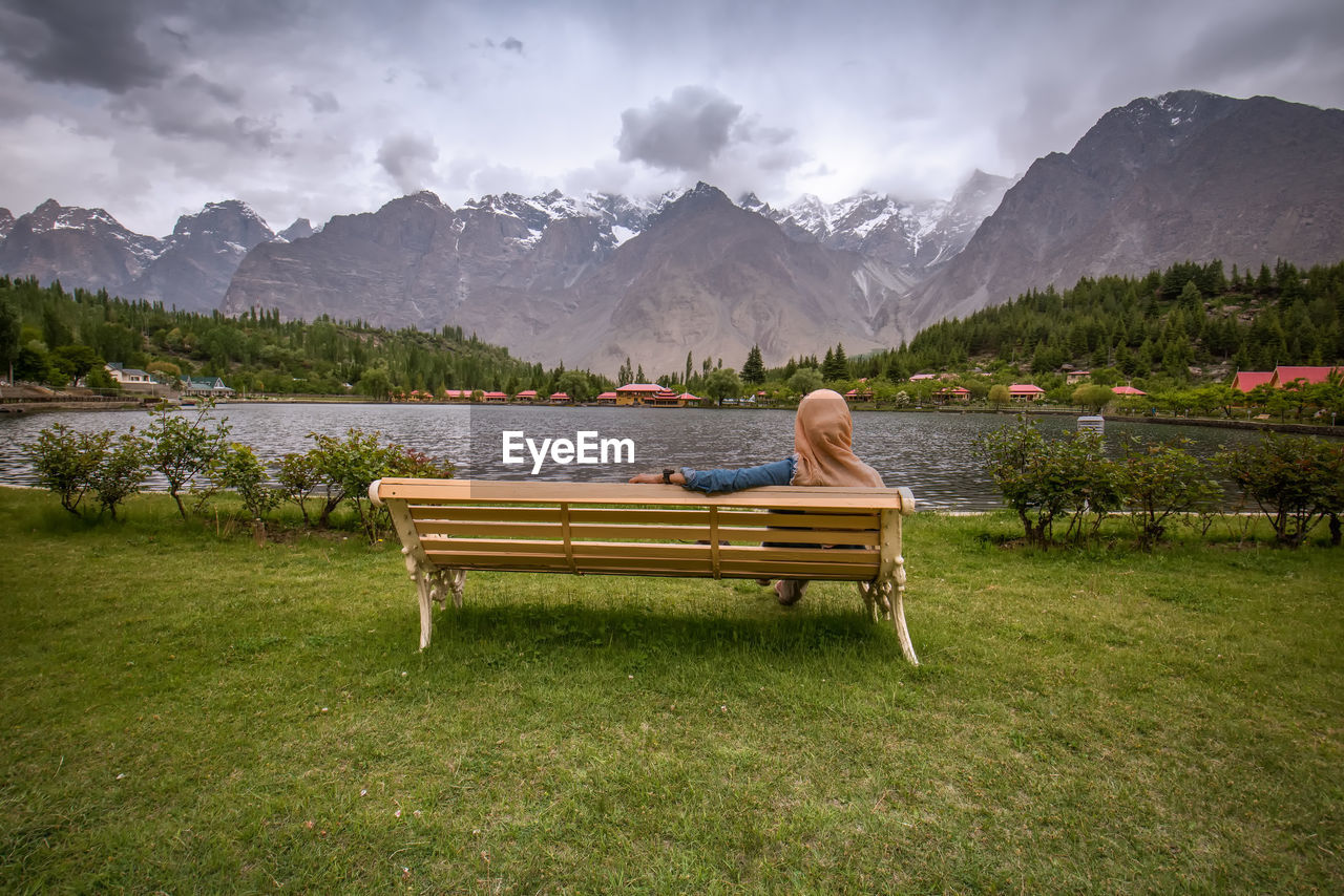 Rear view of woman sitting on bench by lake against mountains