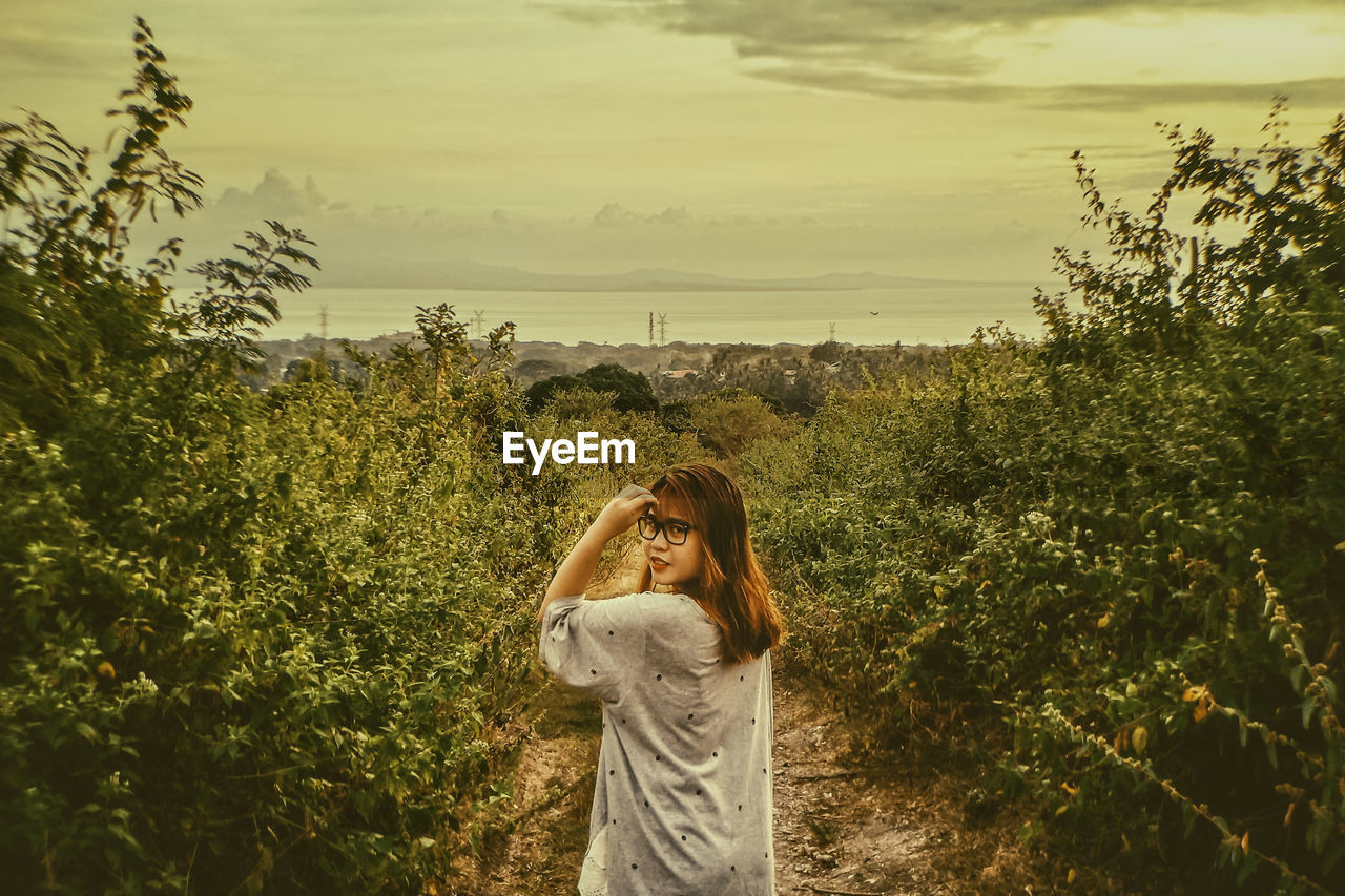 Portrait of young woman standing by plants against sky