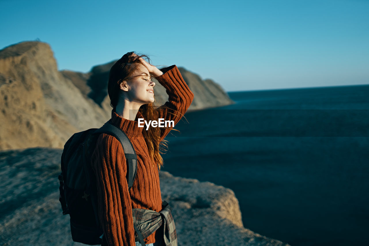 YOUNG WOMAN LOOKING AWAY WHILE STANDING ON ROCK AGAINST SEA