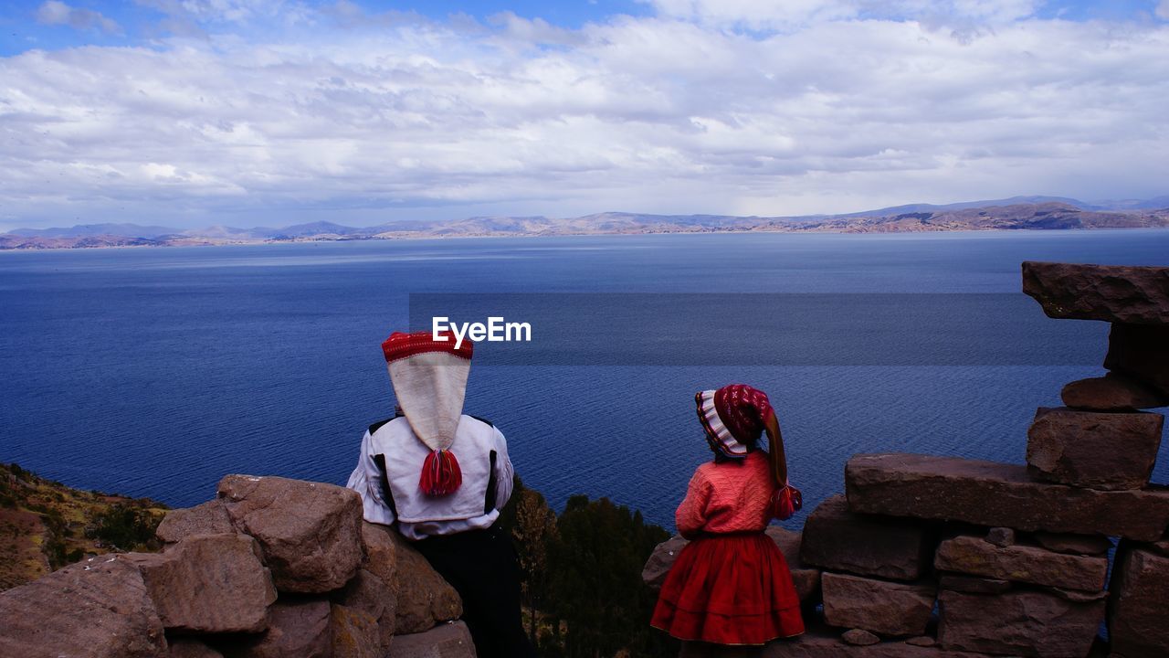 REAR VIEW OF WOMAN LOOKING AT SEA BY MOUNTAINS AGAINST SKY