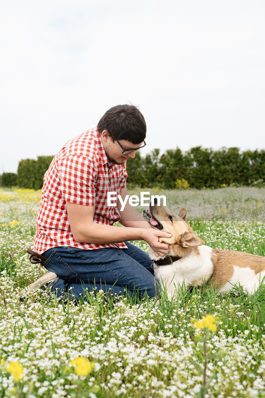 plant, grass, nature, adult, dog, one animal, canine, men, casual clothing, pet, domestic animals, mammal, animal themes, leisure activity, relaxation, animal, sitting, flower, emotion, person, young adult, one person, full length, lifestyles, carnivore, field, day, flowering plant, positive emotion, love, sky, rural scene, lawn, bonding, plain, meadow, landscape, happiness, smiling, outdoors, checked pattern, copy space, clothing, friendship, summer, picnic blanket, food and drink, tranquility, looking, child, enjoyment, women, holding, portrait photography, crouching