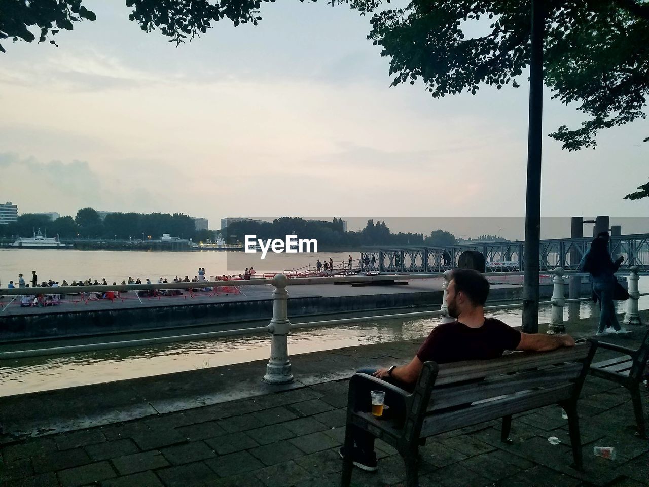 Rear view of mid adult man sitting on bench by river against sky in city during sunset