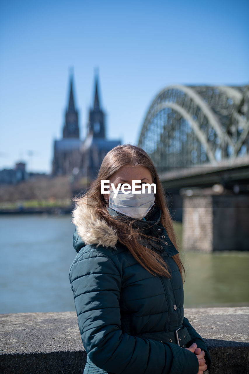 Portrait of young woman wearing mask standing against bridge over river