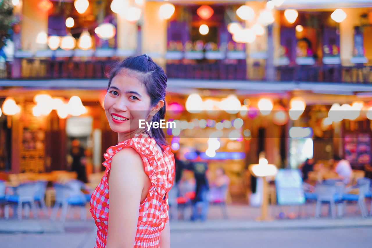 Portrait of smiling woman standing against illuminated restaurant