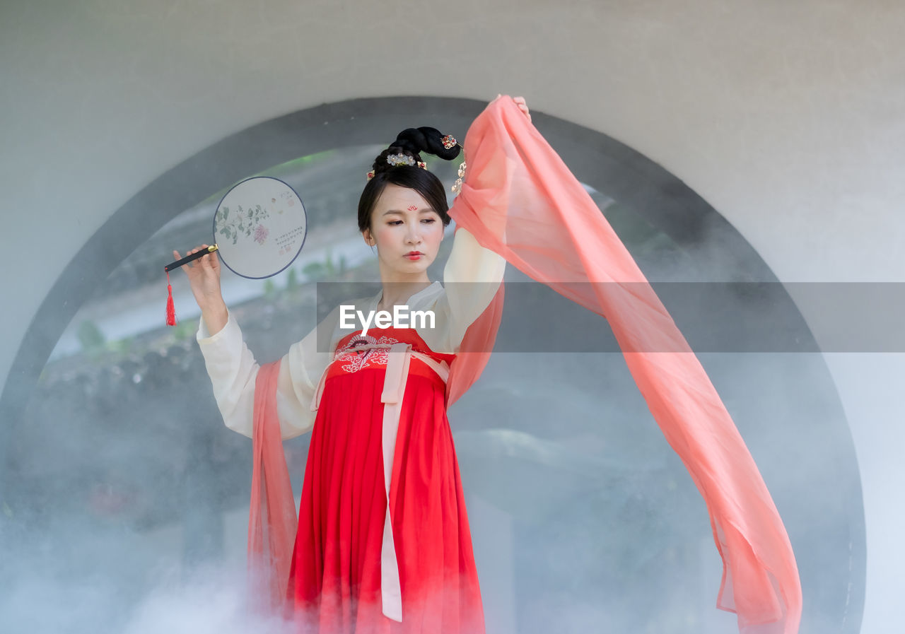 Woman in traditional clothing holding hand fan against window