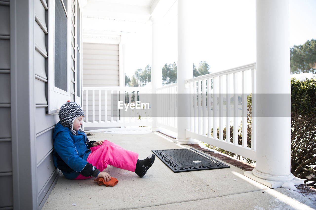 Wide view of boy in pink snow pants sitting on snowy front porch