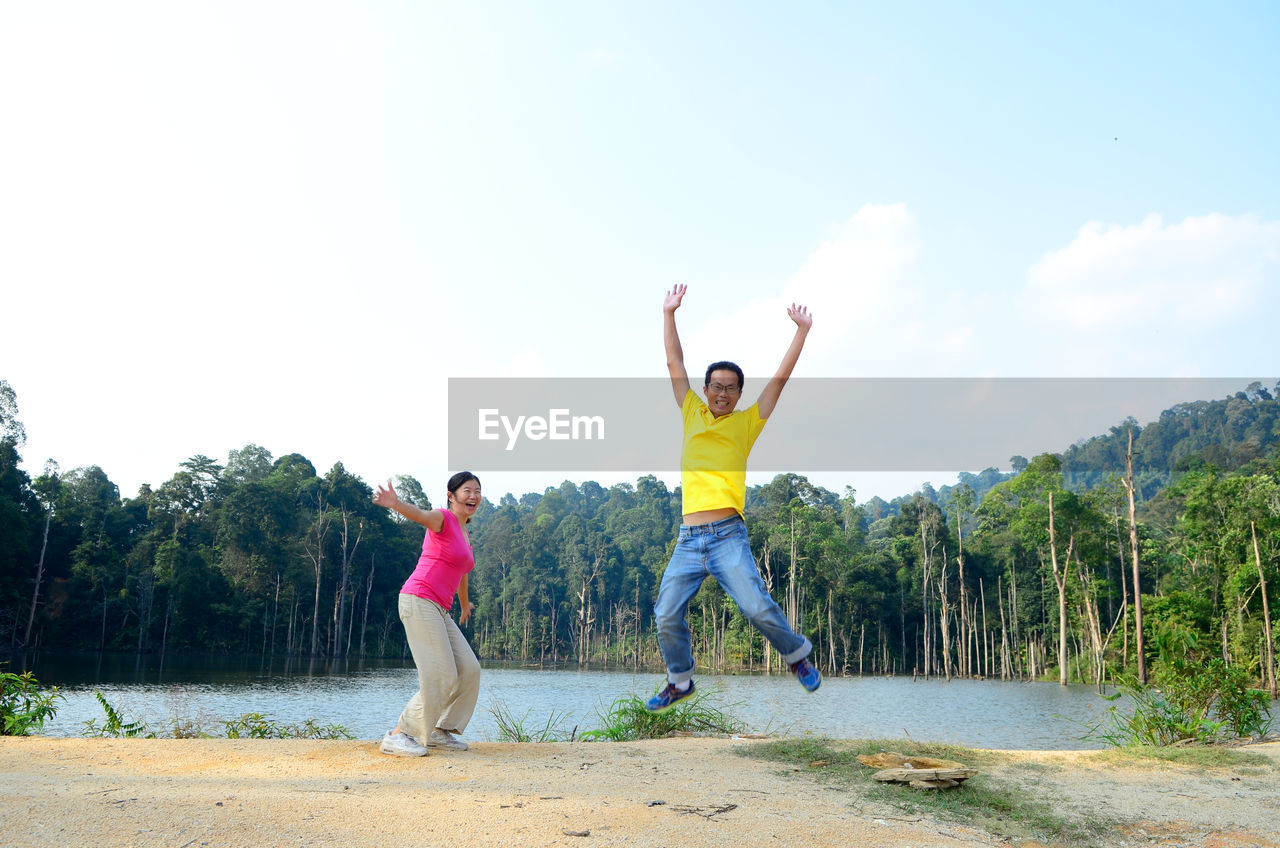 Cheerful people with arms outstretched jumping by lake against forest and sky