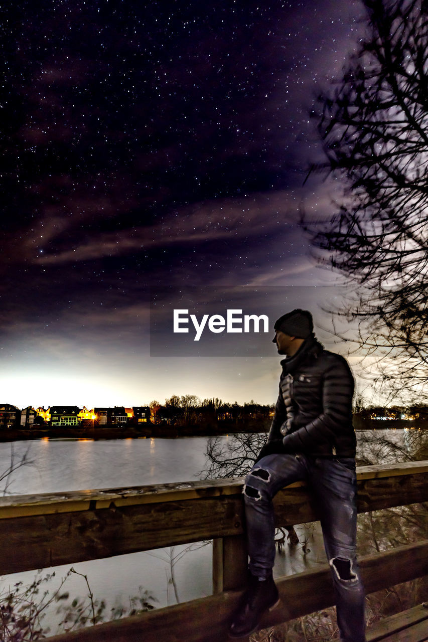 Man sitting on railing by lake against star field