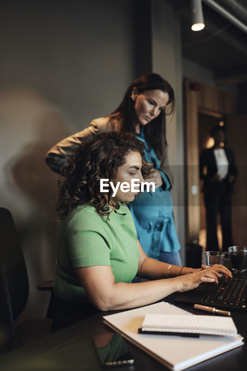 Businesswoman working on laptop by female pregnant colleague in office