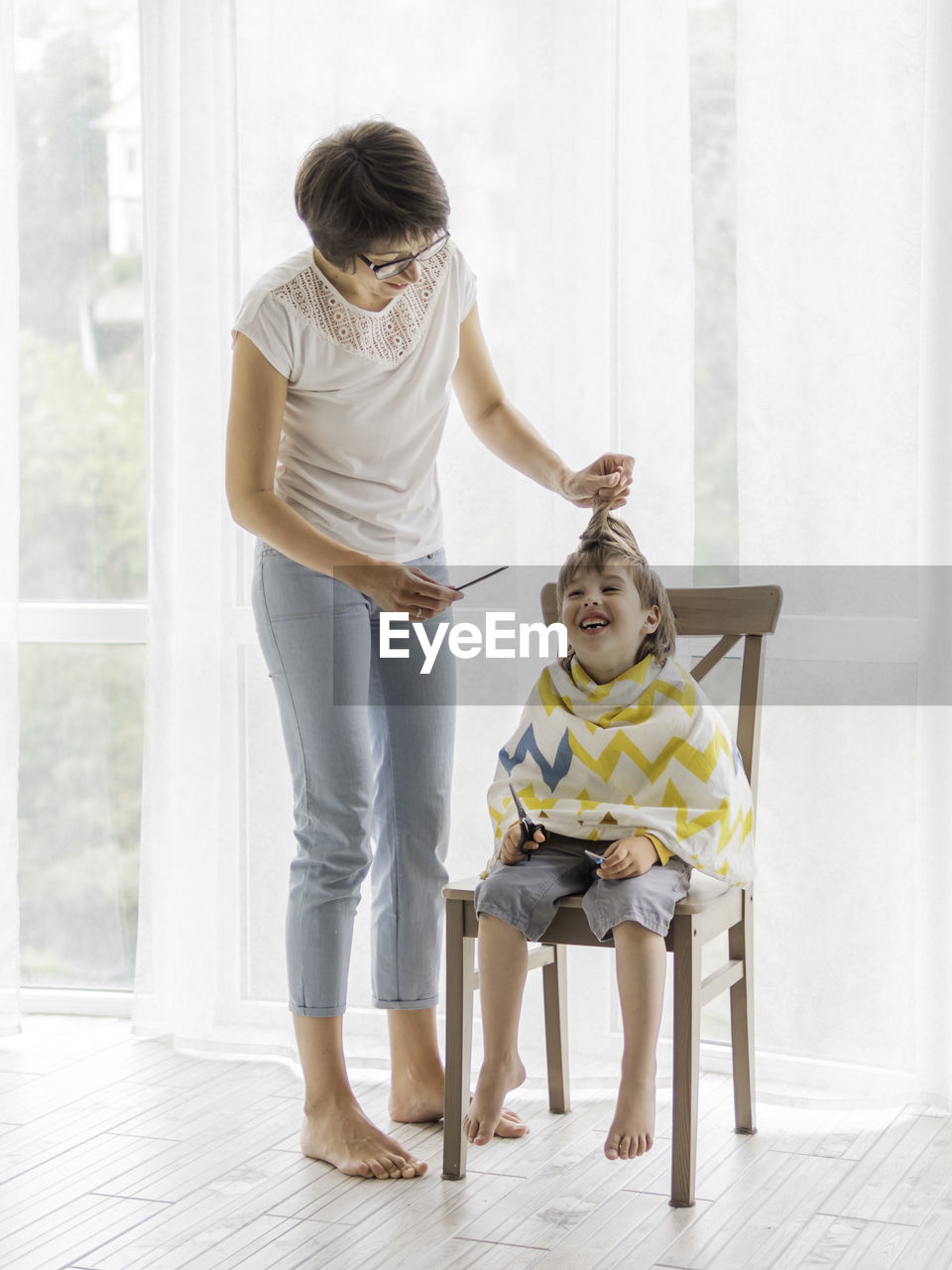 Mother cuts her son's hair by herself. little boy sits, covered with cloth, holds pair of scissors. 