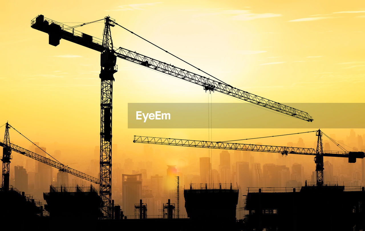 LOW ANGLE VIEW OF CRANES AT CONSTRUCTION SITE AGAINST SKY DURING SUNSET