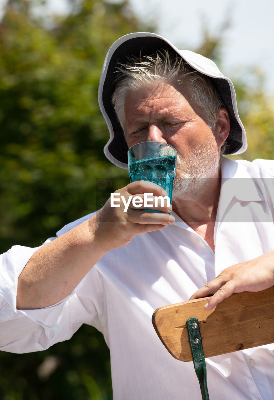 An elderly man sits outside in the great heat. he sweats and drinks mineral water.