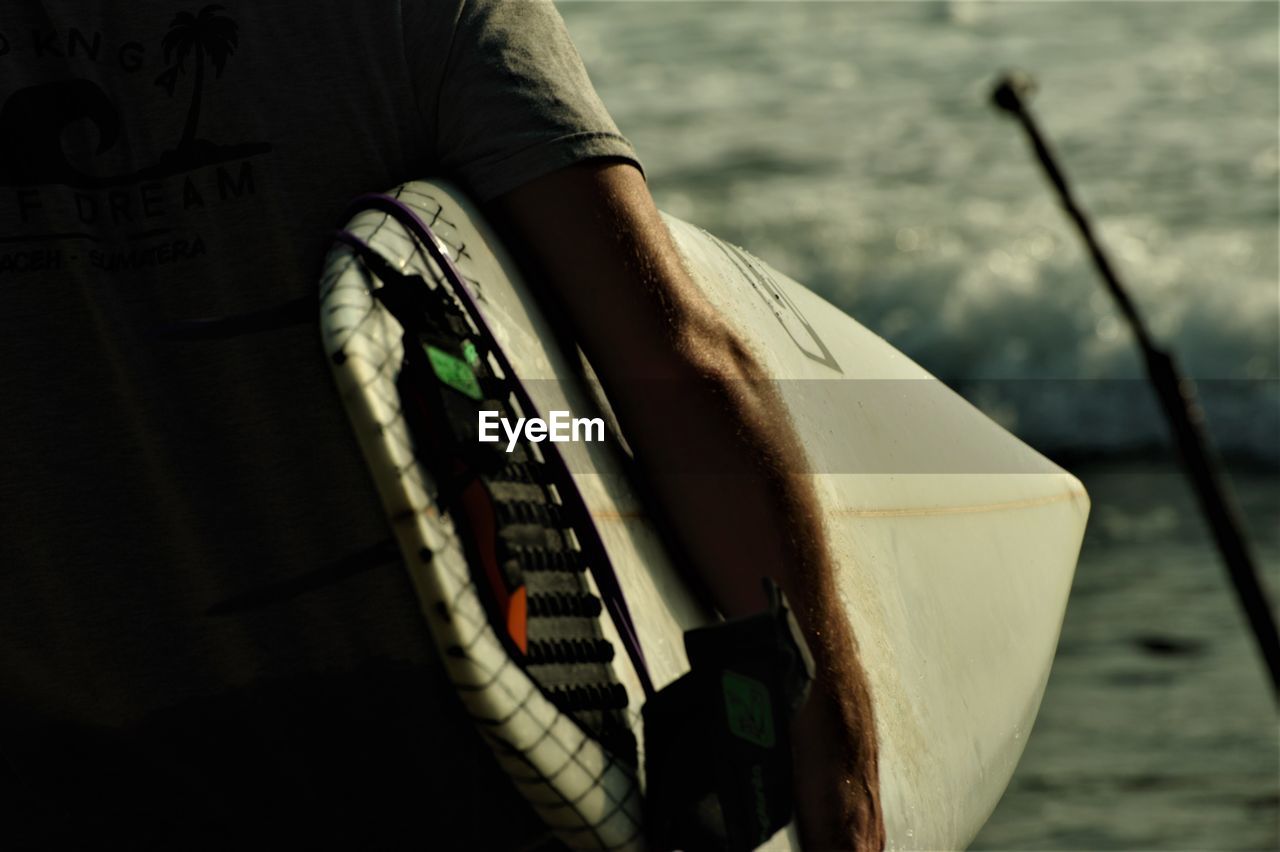 Close-up of man carrying surfboard at beach during sunset