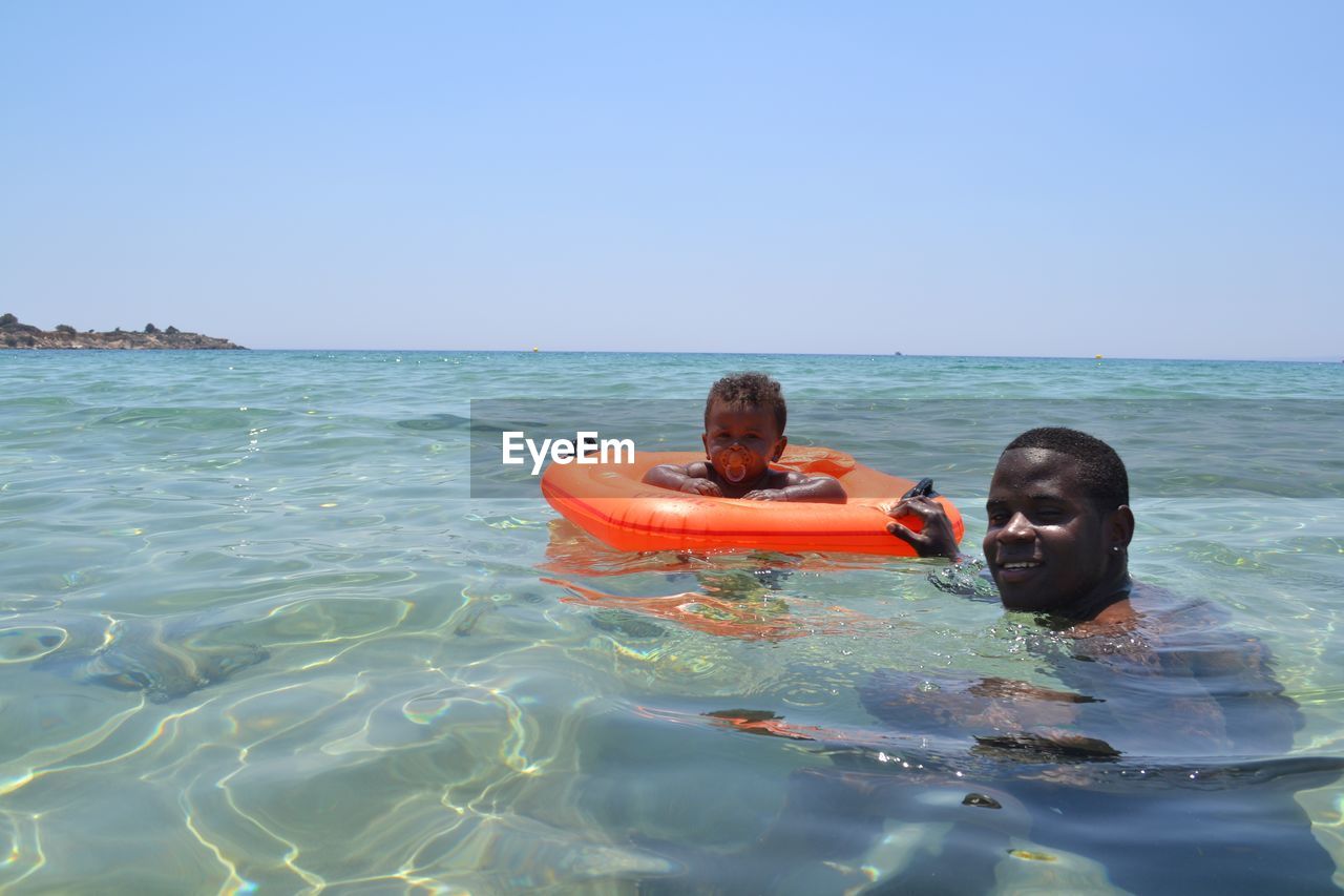 Portrait of man with baby wearing inflatable ring while swimming in sea against clear sky during sunny day