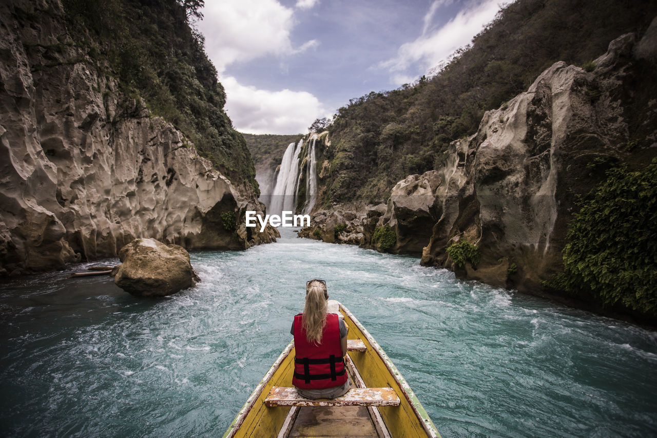 Rear view of woman sitting in boat on river by mountains against waterfall