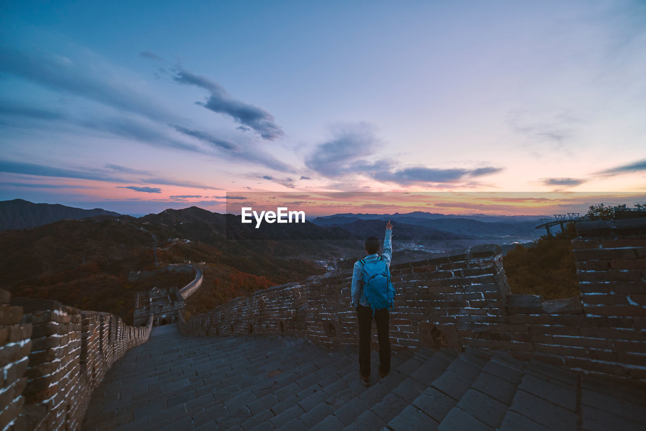 Rear view of mature man with backpack standing on steps against sky during sunset