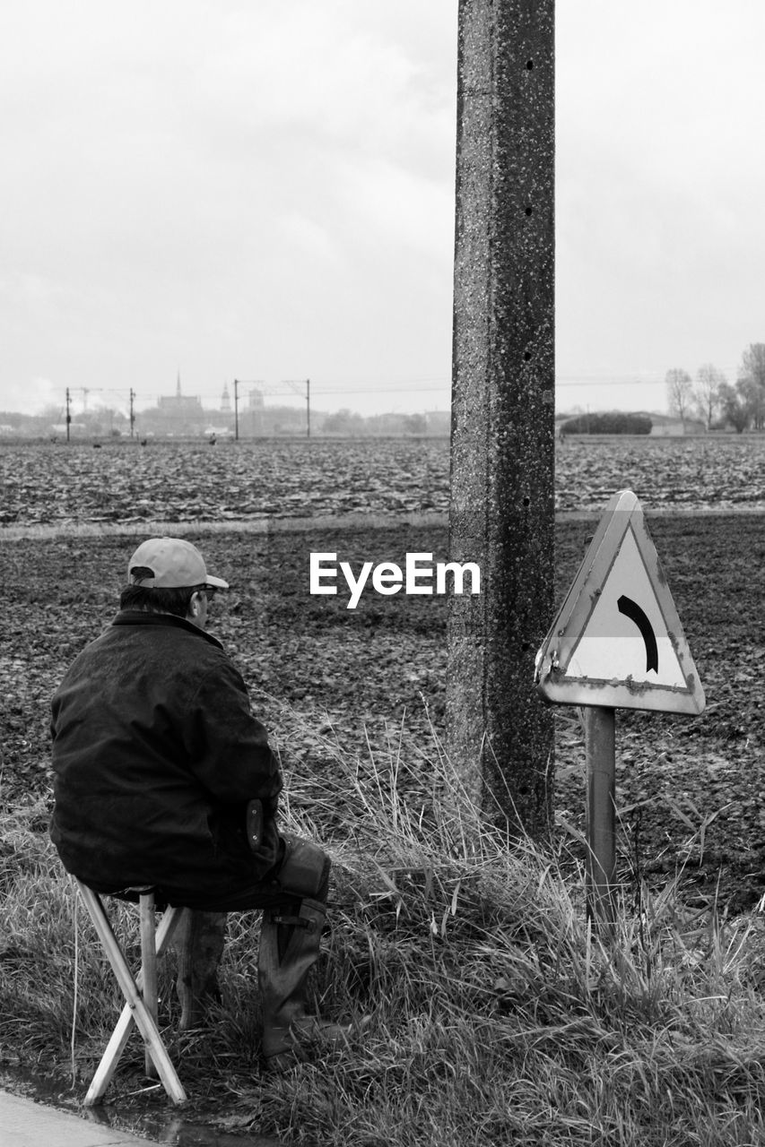 REAR VIEW OF MAN SITTING ON FIELD BY ROAD