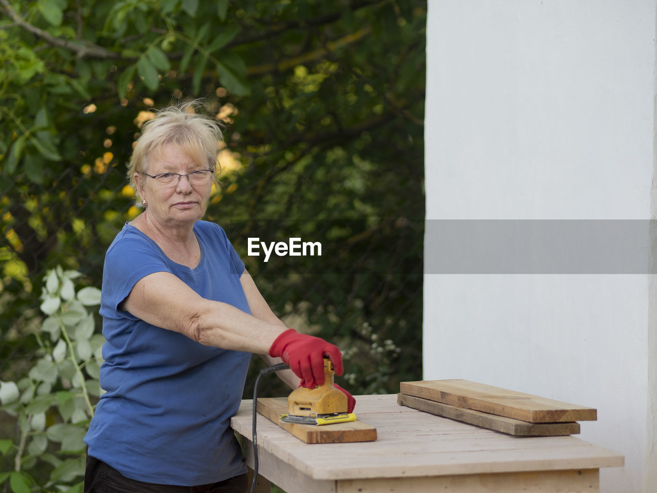 Senior caucasian woman in a blue t-shirt and red gloves is sanding a board with a sander 