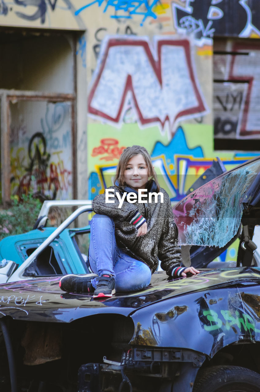 PORTRAIT OF SMILING YOUNG WOMAN SITTING ON CAR