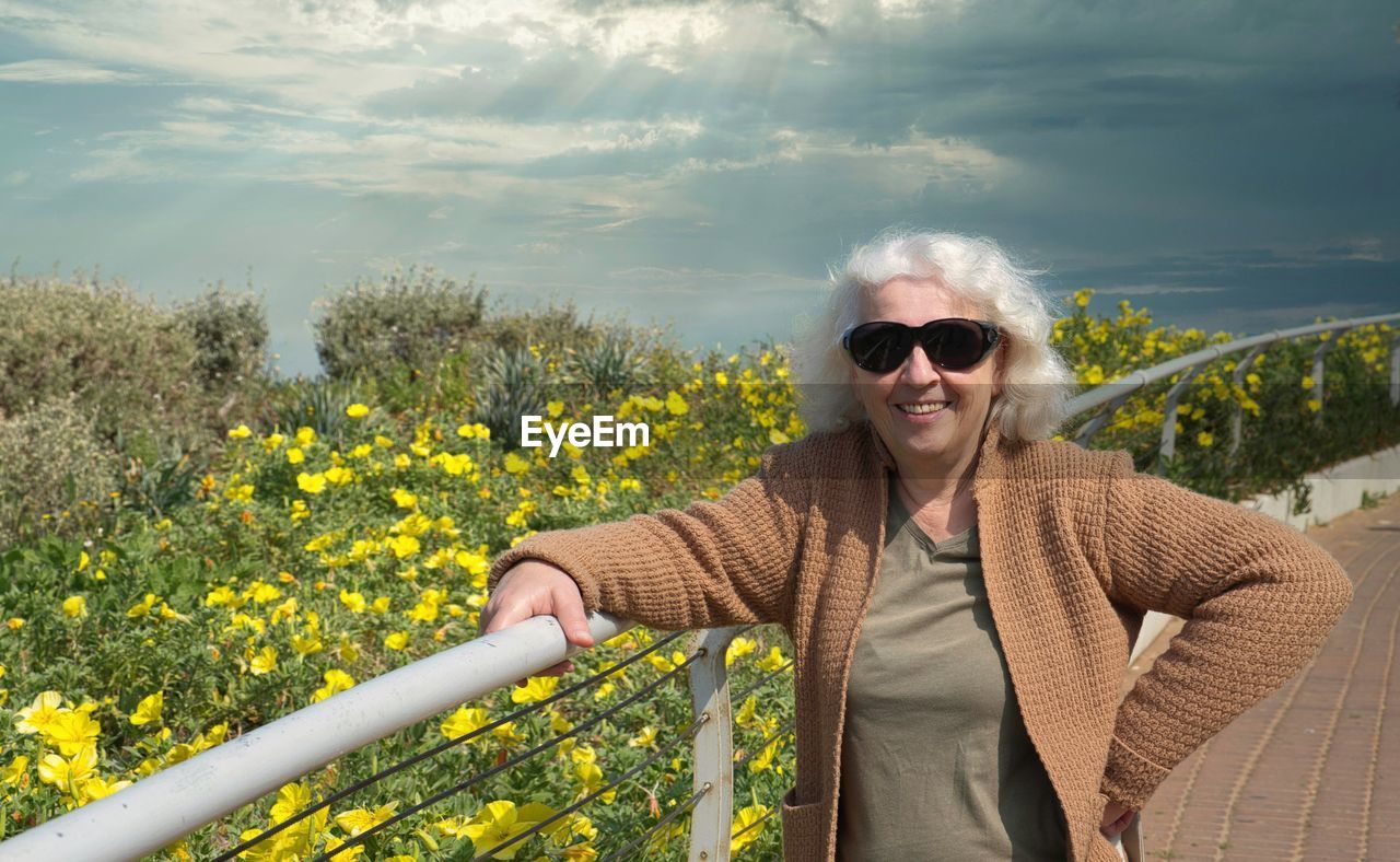 Mature gray-haired happy woman over 60, wearing sunglasses, against the background of yellow flower