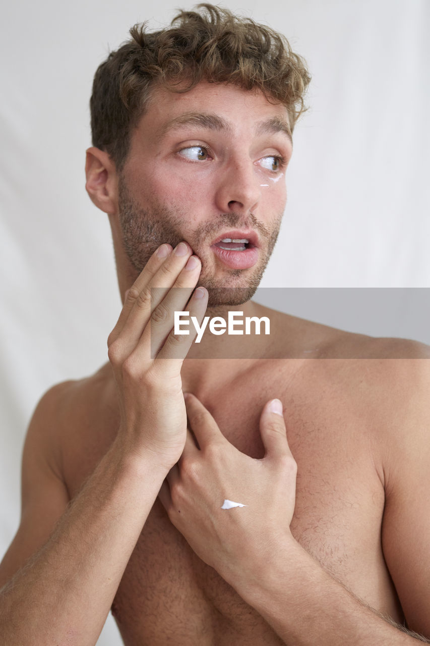 Young man applying moisturizer while looking away over white background