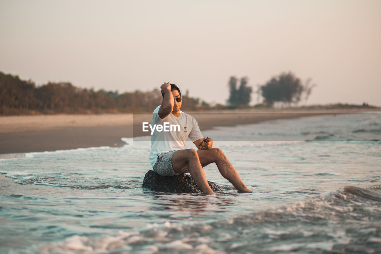 Side view of good looking young man sitting on a rock at the beach with feet in water