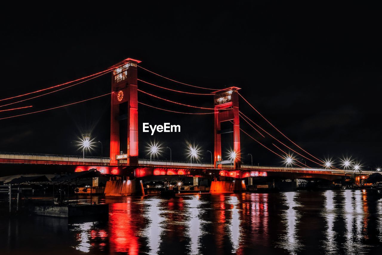 Illuminated suspension bridge over river against sky at night