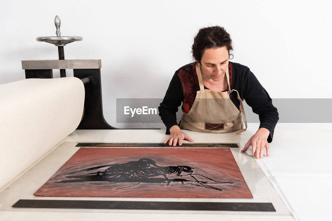 Focused middle aged female master putting sheet of linoleum with inked ornament on printing press while working in linocut technique in work studio