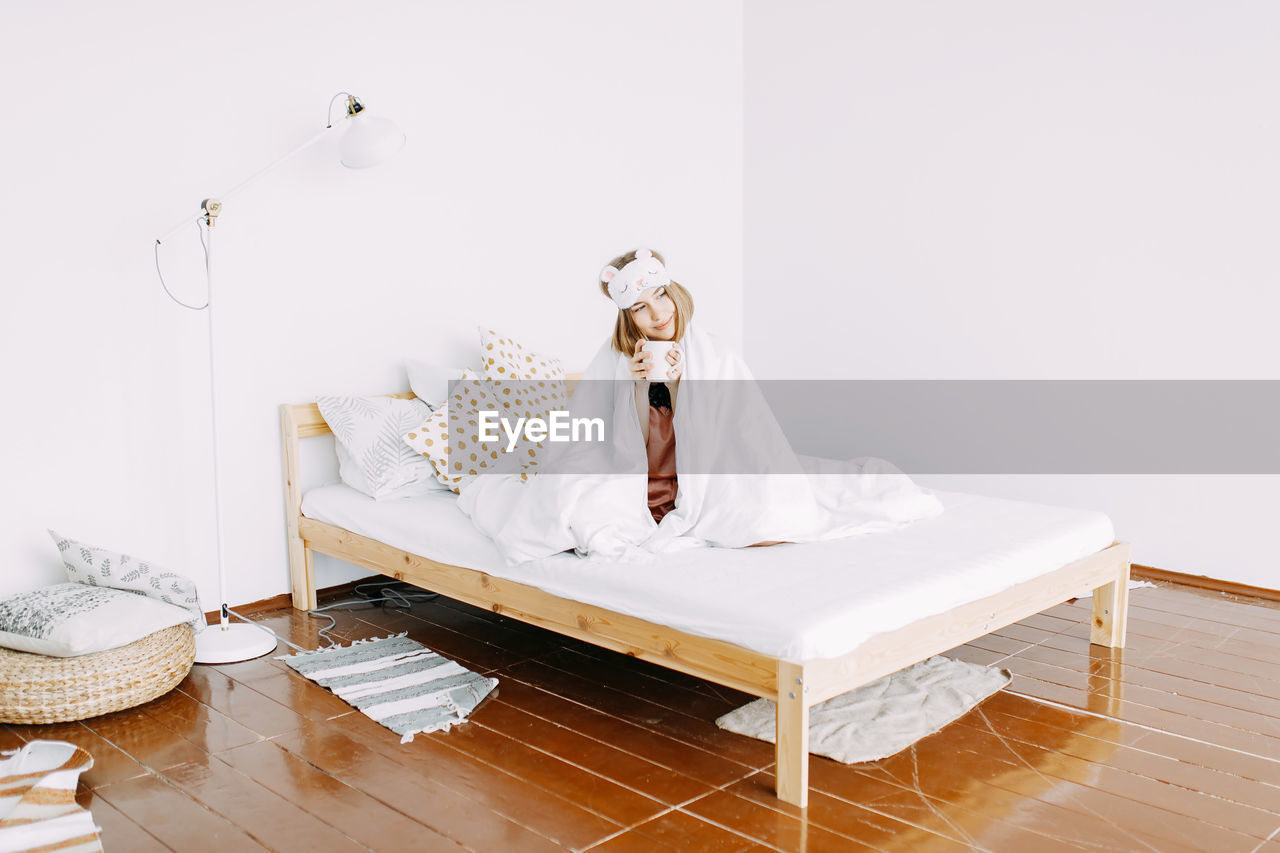 A young sick woman in pink pajamas and a sleeping mask on her head is sitting on the bed