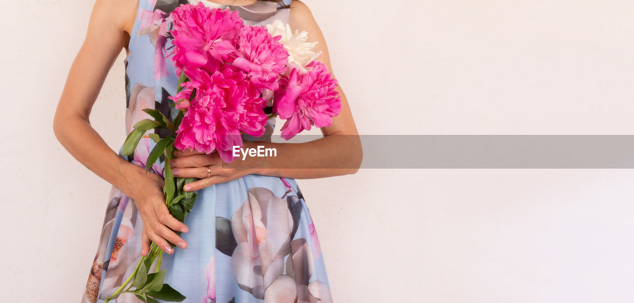MIDSECTION OF WOMAN HOLDING PINK FLOWERS AGAINST WALL