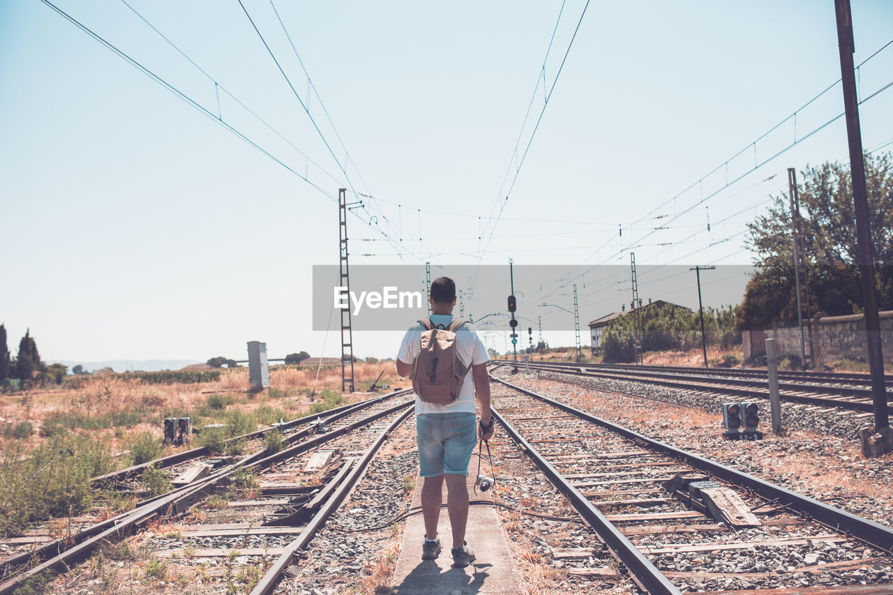 Rear view of man standing on railroad tracks against sky during sunny day