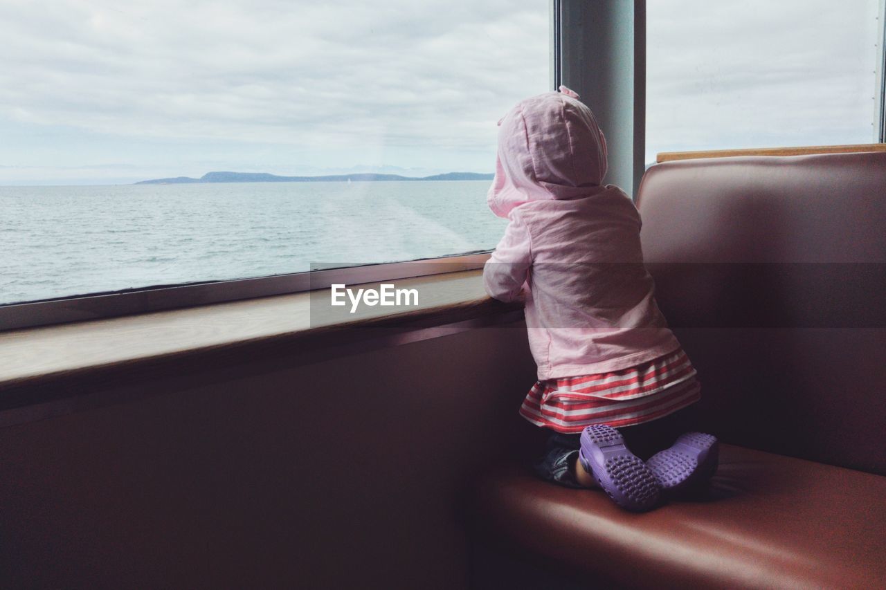 Rear view of girl looking through ferry window while wearing hooded shirt