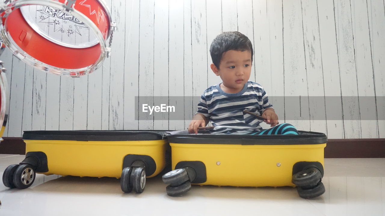 Cute boy sitting in luggage by drums at home