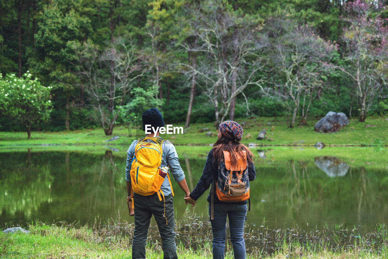Rear view of couple holding hands while standing by lake at forest