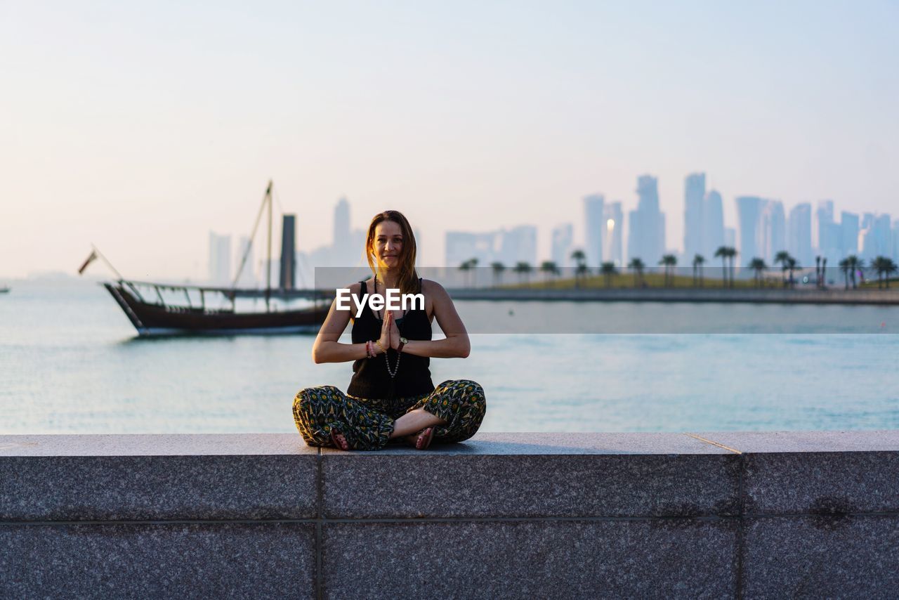 YOUNG WOMAN SITTING ON SEA AGAINST SKY IN CITY