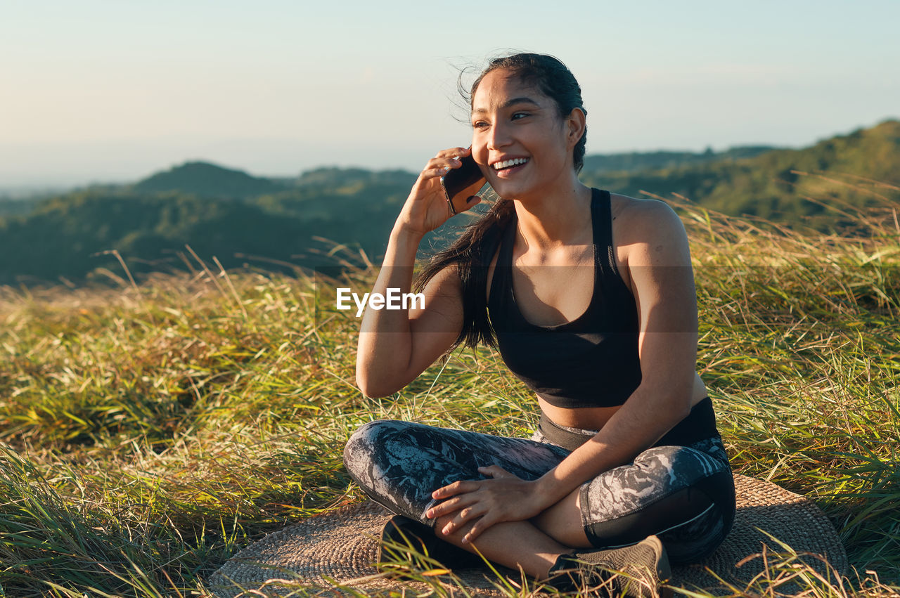 Young woman talking on phone while sitting against landscape