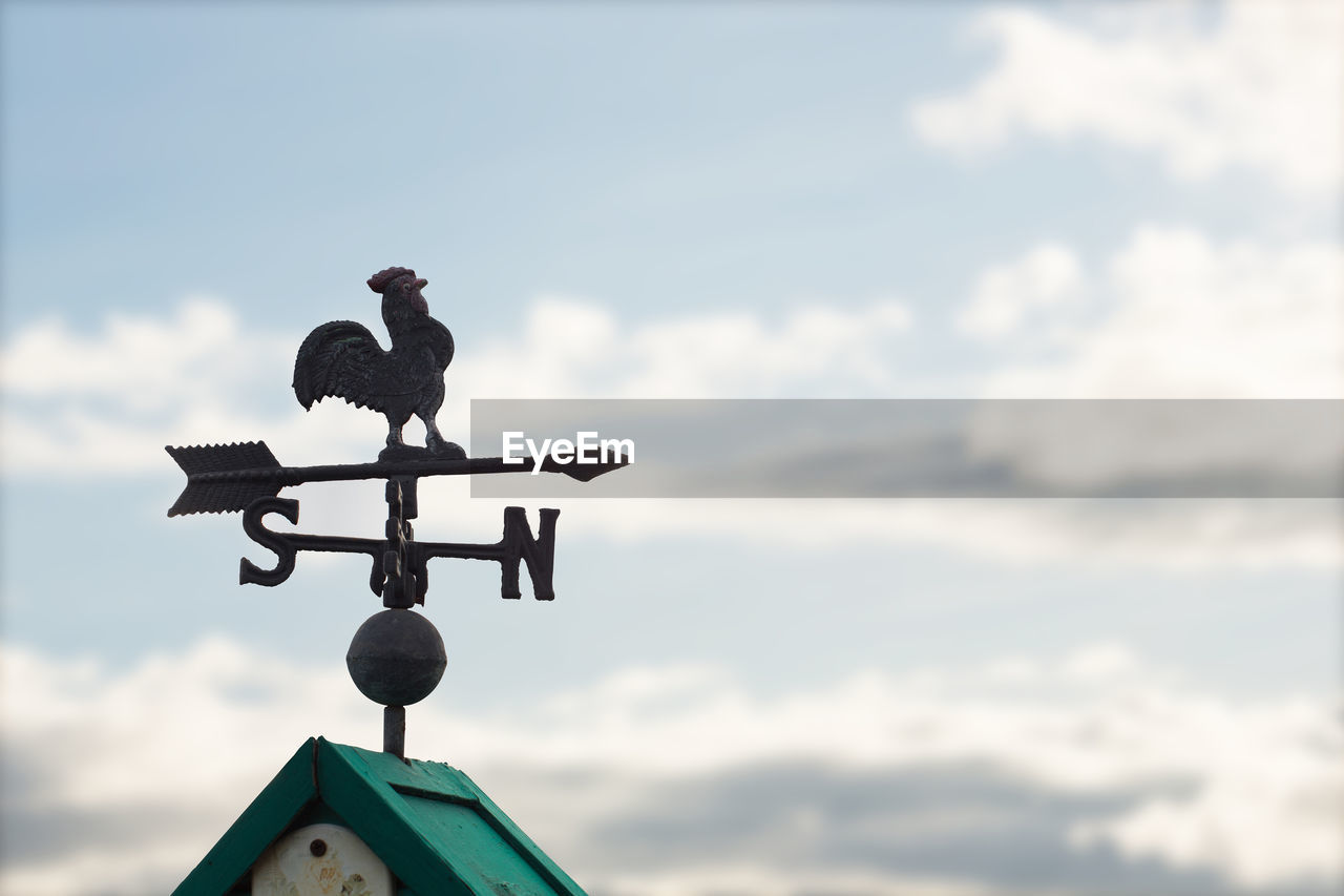 Weather vane on rooftop against cloudy sky