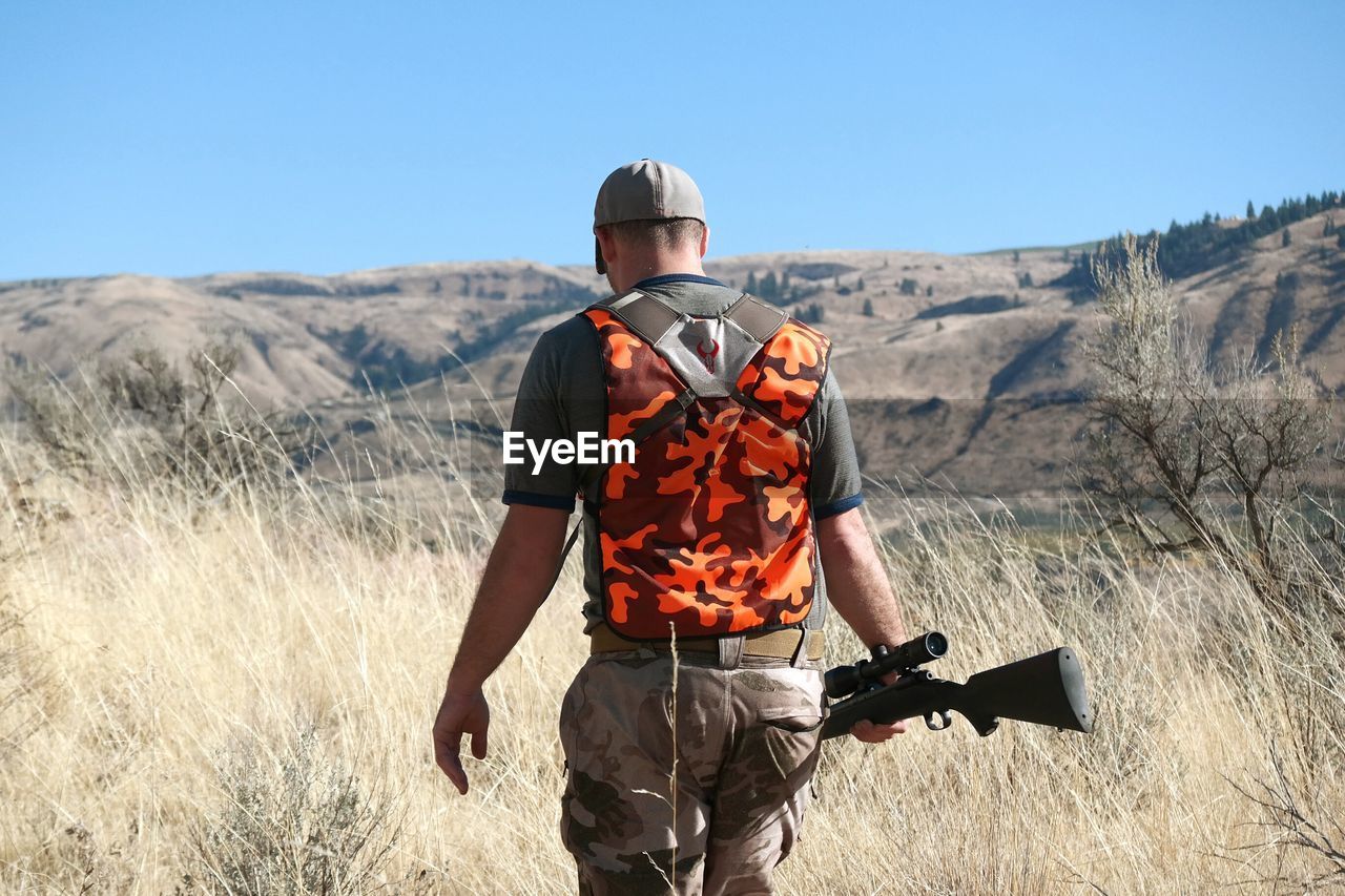 Rear view of man with sniper standing on field against clear sky
